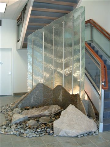 Law firm Olympia Wa. Lobby new construction. 12 ft x 12 ft tall presentation of glass, stainless steel and stone, hand made glass panels set up at a 90 degree corner radius, stainless steel verticals add support and water delivery, Idaho quarts MT. Range front and back, beach rock covering sunk in hidden basin, up lighting within the MT. range for a dramatic affect.
